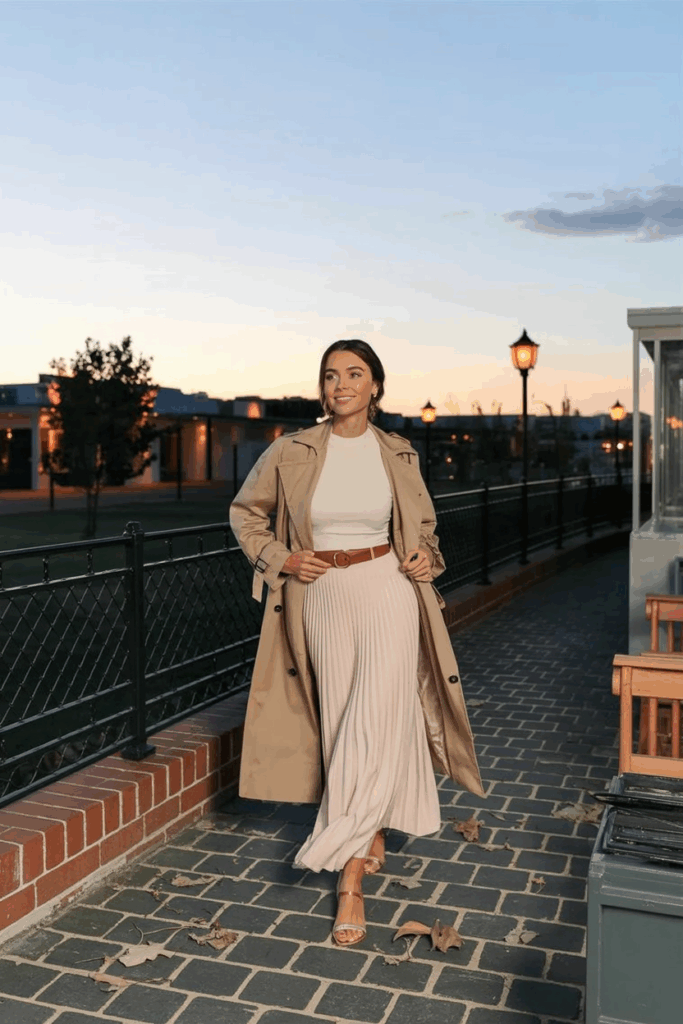 A woman wearing a pale blush modest maxi skirt with a fitted ivory top and a light tan trench coat, belted at the waist