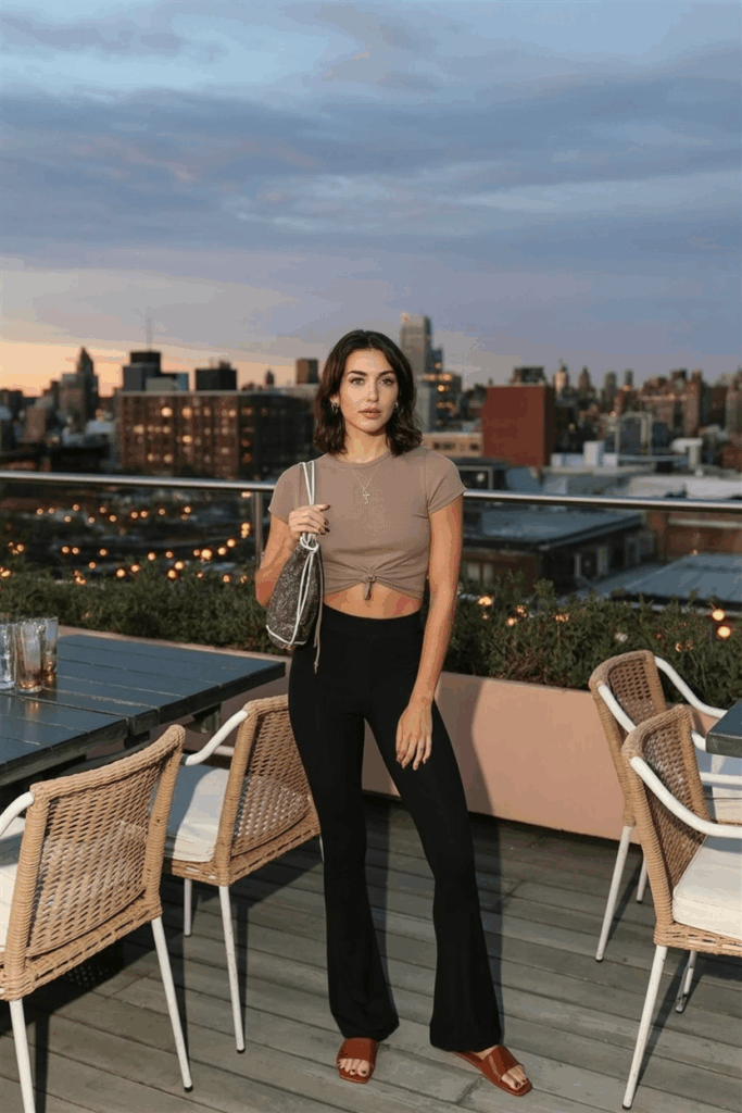 A woman in black flare leggings, a cropped sand-colored boxy tee, brown leather slides, and a drawstring pouch bag.