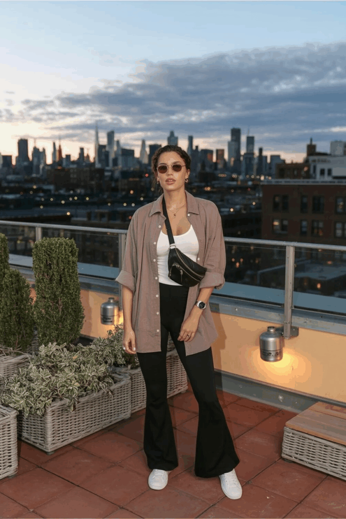 A woman in black flare leggings, a taupe linen button-up shirt layered over a white tank, white leather sneakers, and a crossbody belt bag. She wears aviator sunglasses and a watch