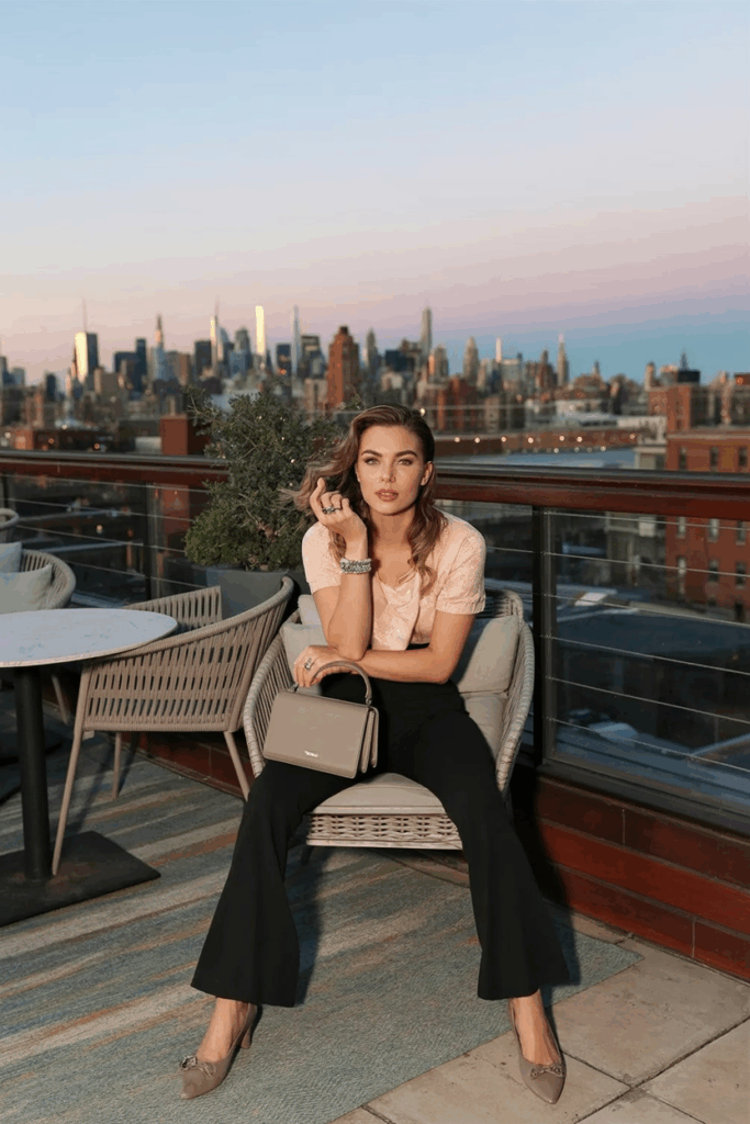 A woman seated in black flare leggings, a soft knit top in blush pink, taupe ballet flats, and a structured mini handbag. Her hair is in loose waves and she wears a dainty ring stack