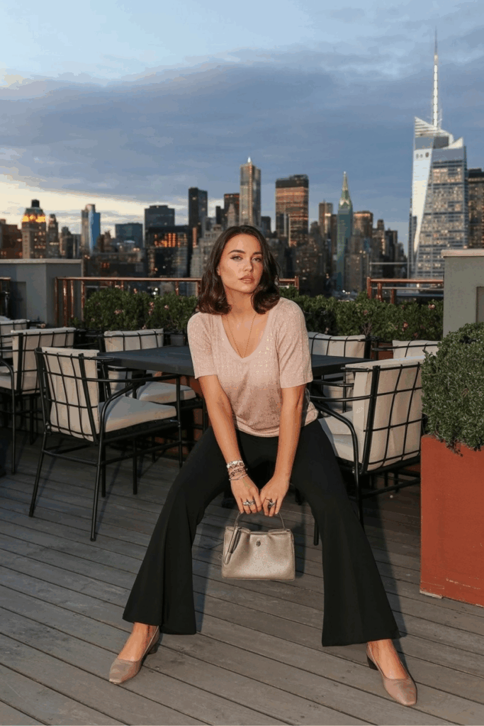 A woman seated in black flare leggings, a soft knit top in blush pink, taupe ballet flats, and a structured mini handbag. Her hair is in loose waves and she wears a dainty ring stack