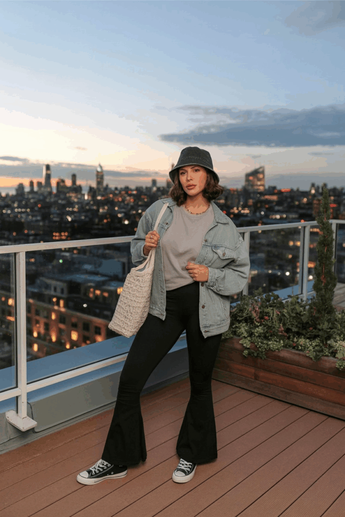 A woman styled in black flare leggings, an oversized light-gray tee, a light-wash denim jacket, and classic white Converse. She wears a bucket hat and carries a reusable tote