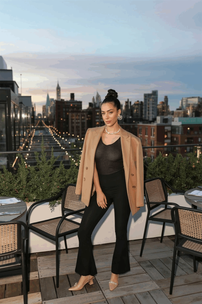 woman wearing black flare leggings, a slim black knit top, a camel blazer, and nude slingback flats. She has her hair in a sleek bun and wears small pearl earrings