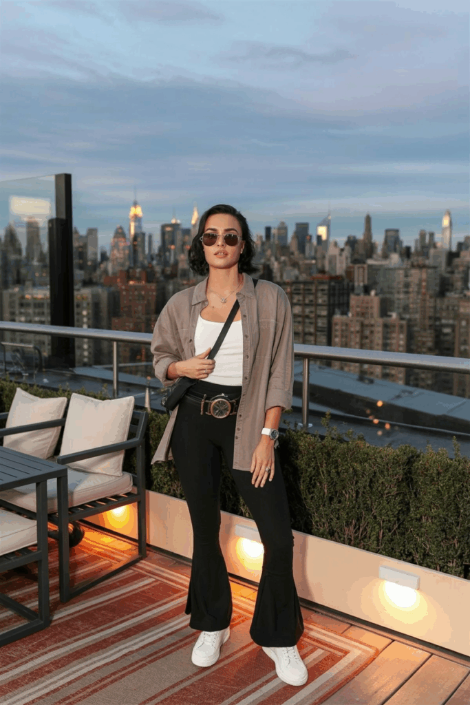 A woman in black flare leggings, a taupe linen button-up shirt layered over a white tank, white leather sneakers, and a crossbody belt bag. She wears aviator sunglasses and a watch