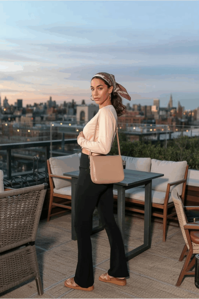 A woman wearing black flare leggings, a soft cream long-sleeve tee, tan leather sandals, and a beige crossbody bag. A printed silk bandana is tied into her low ponytail. She wears thin gold hoop earrings