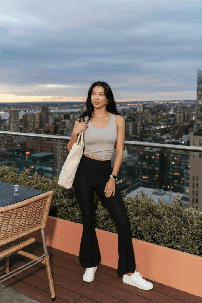 woman in sleek, high-waisted black flare leggings, a tucked-in ivory seamless tank top, white low-top sneakers, and small gold hoops. She carries a canvas tote bag and wears a digital watch