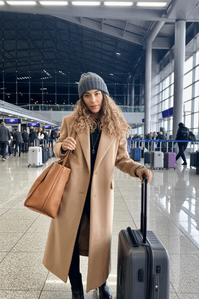 woman adjusting a gray wool beanie over soft waves, wearing a long camel coat and holding a tan leather tote