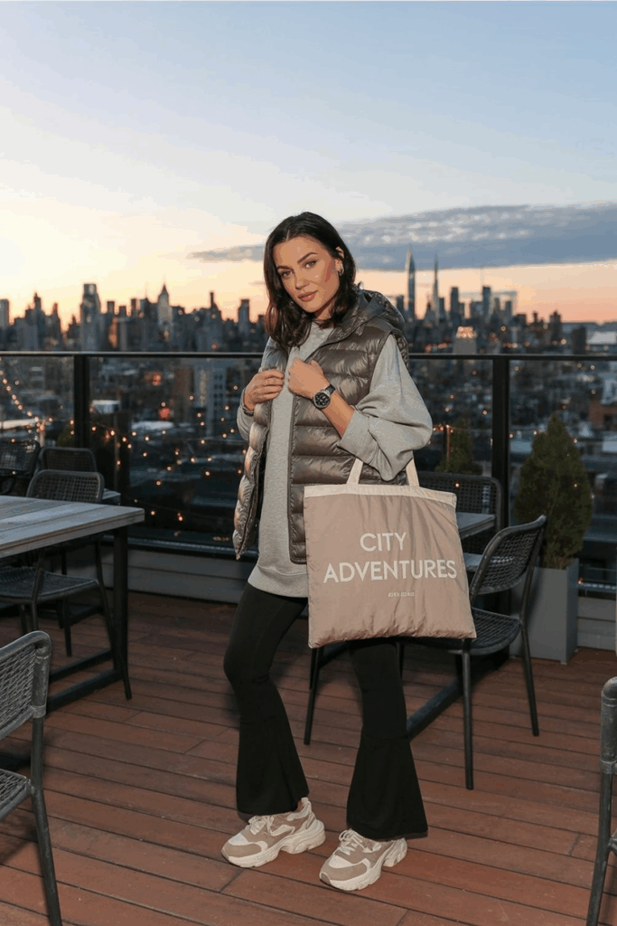 A woman in black flare leggings paired with beige and white chunky sneakers, a relaxed oversized sweatshirt, and a puffer vest. She carries a canvas tote and wears a sporty watch.