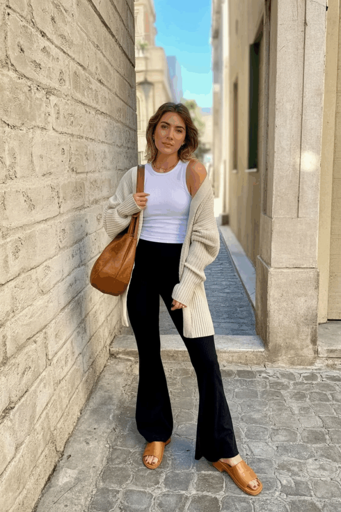 woman in black flare leggings, a fitted white ribbed tank, and a chunky oatmeal cardigan. She wears tan leather mules and carries a slouchy hobo bag