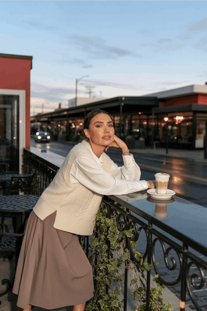 A woman wearing a cream knit vest over a long-sleeve white blouse, tucked into a muted brown skirt.