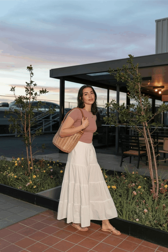 A woman wearing a soft white tiered maxi skirt made of lightweight cotton, styled with a slim-fit dusty rose top. She’s holding a woven basket bag