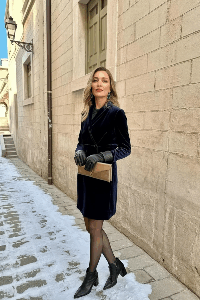 A woman wearing a black velvet dress, black wool-blend tights, emerald earrings, a bronze clutch, leather gloves, and black heeled boot