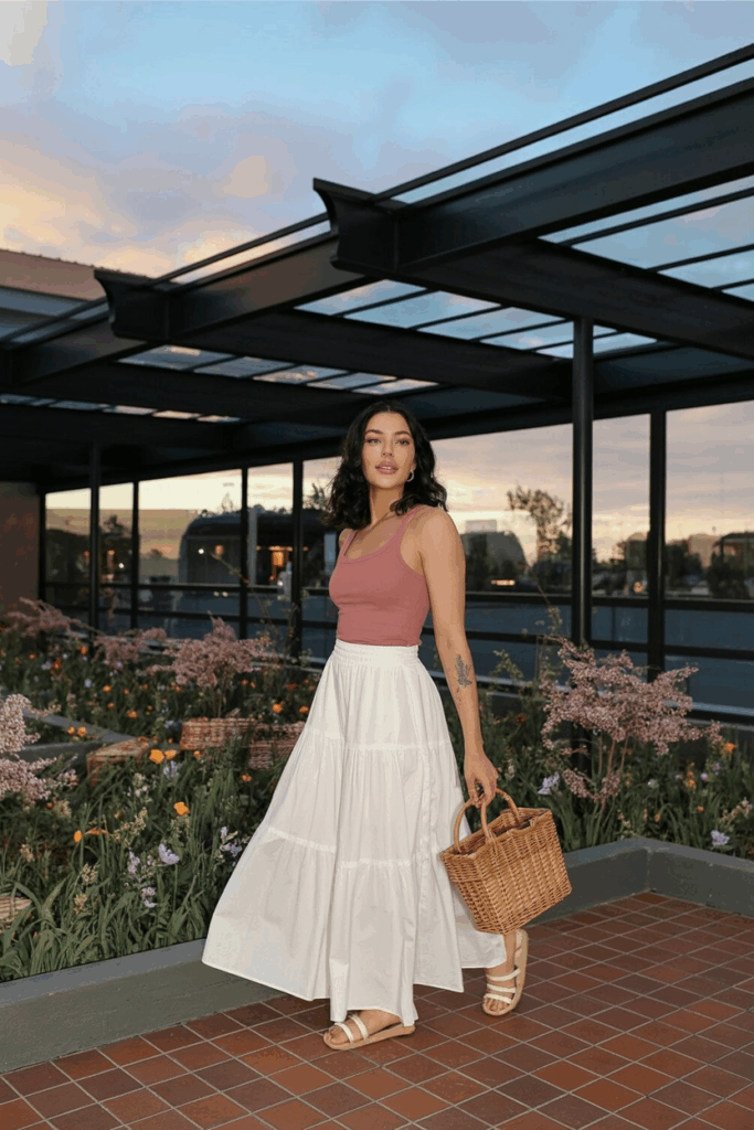 A woman wearing a soft white tiered maxi skirt made of lightweight cotton, styled with a slim-fit dusty rose top. She’s holding a woven basket bag