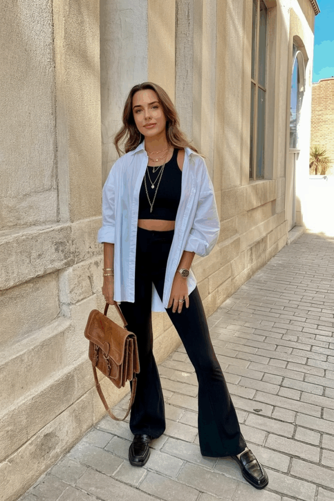 A woman in black flare leggings and an oversized white button-up shirt, loosely tucked in front. She wears black loafers, layered gold necklaces, and carries a leather satchel