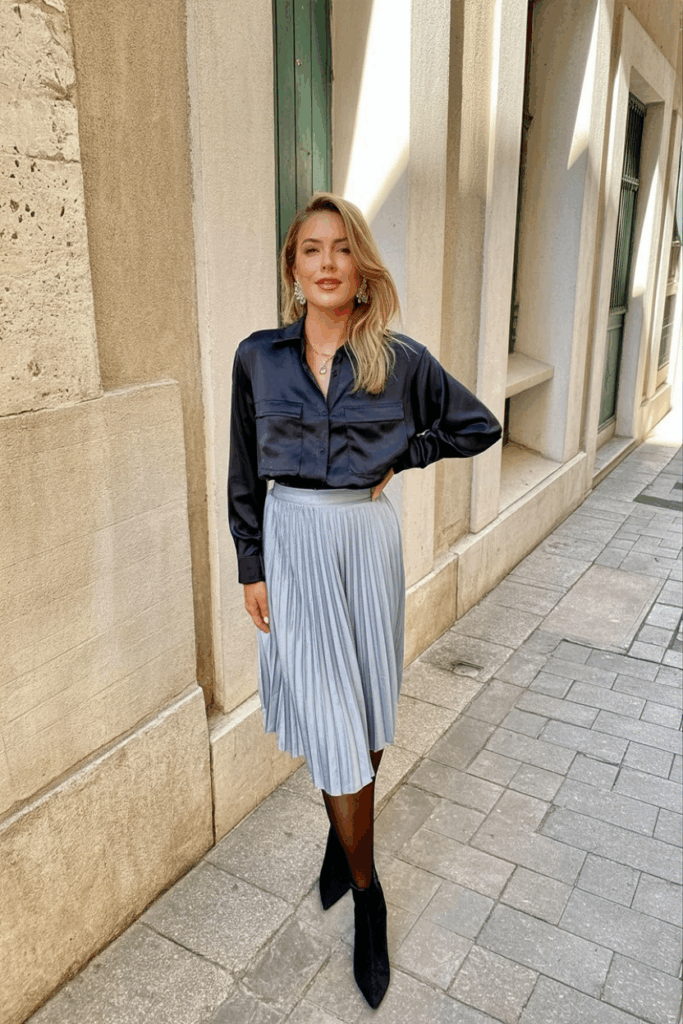woman wearing a midnight-blue satin blouse and matching pleated skirt, black cashmere blend tights, paired with silver statement earrings and black pointed ankle boots