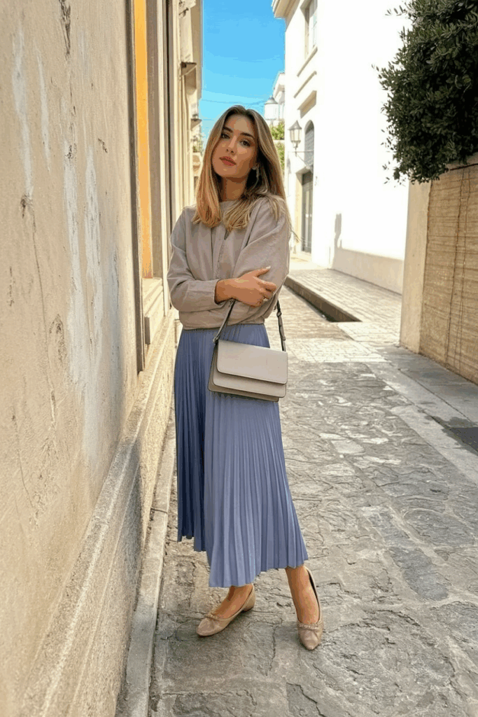 A woman wearing a dusty lavender pleated skirt and nude ballet flats. A soft crossbody bag at her side.