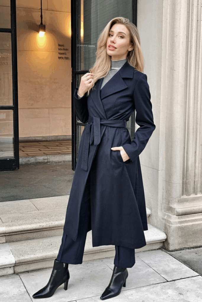 A woman wearing a navy wrap coat belted at the waist over a striped turtleneck and tailored trousers. She carries a wool tote and wears low-heeled boots