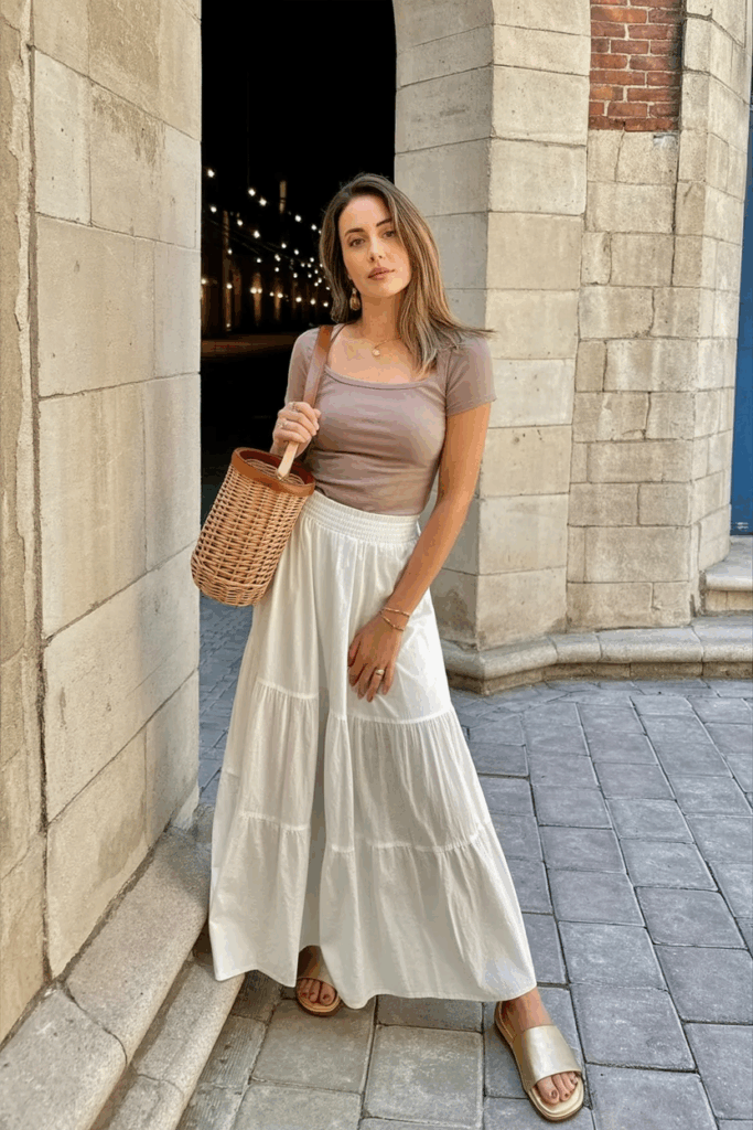 A woman wearing a soft white tiered maxi skirt made of lightweight cotton, styled with a slim-fit dusty rose top. She’s holding a woven basket bag