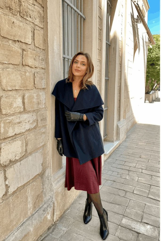 woman wearing a tailored navy wool wrap coat over a burgundy midi dress, black opaque tights, leather gloves, and black heeled booties