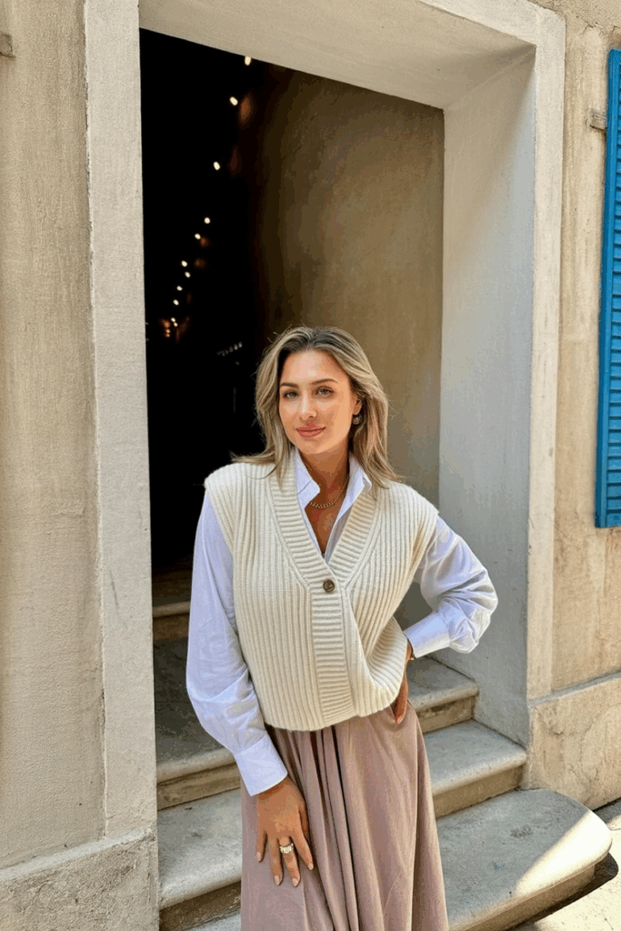 A woman wearing a cream knit vest over a long-sleeve white blouse, tucked into a muted brown skirt.