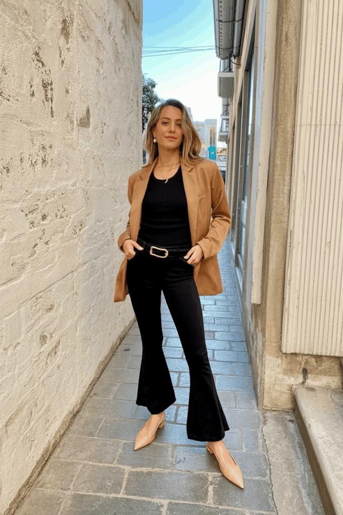 woman wearing black flare leggings, a slim black knit top, a camel blazer, and nude slingback flats. She has her hair in a sleek bun and wears small pearl earrings