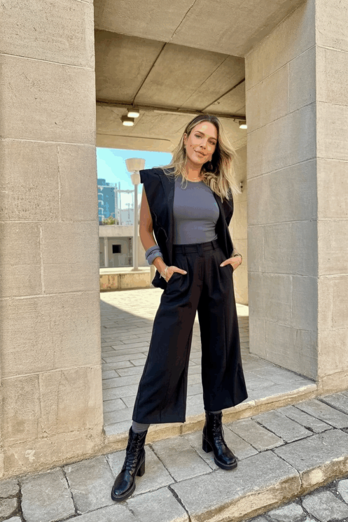 A woman wearing a midnight-black wide-leg jumpsuit with structured shoulders over a fitted thermal top and insulated leggings, with black heeled combat boot