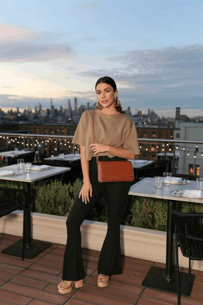 A woman in black flare leggings, a lightweight tan tee, a chestnut leather crossbody bag, gold huggie earrings, and platform sandals. Her nails are neatly polished in nude.