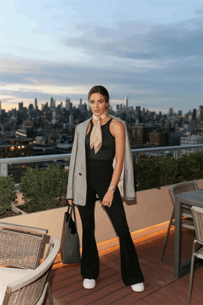 Woman in black flare leggings, a fitted black ribbed tank, white sneakers, and a linen blazer draped over her shoulders. A soft blush-colored lightweight scarf is knotted at her neck. She carries a structured tote and wears a dainty gold bracele
