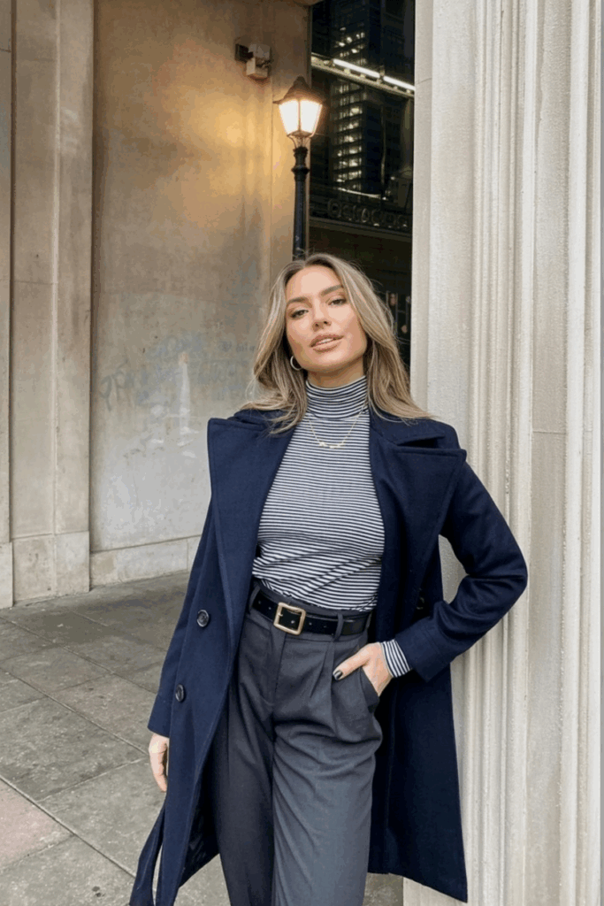 A woman wearing a navy wrap coat belted at the waist over a striped turtleneck and tailored trousers. She carries a wool tote and wears low-heeled boots
