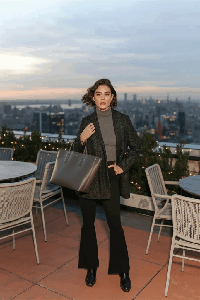 A woman dressed in black flare leggings, a charcoal turtleneck sweater, a dark gray wool coat, and black Chelsea boots. She carries a structured leather tote