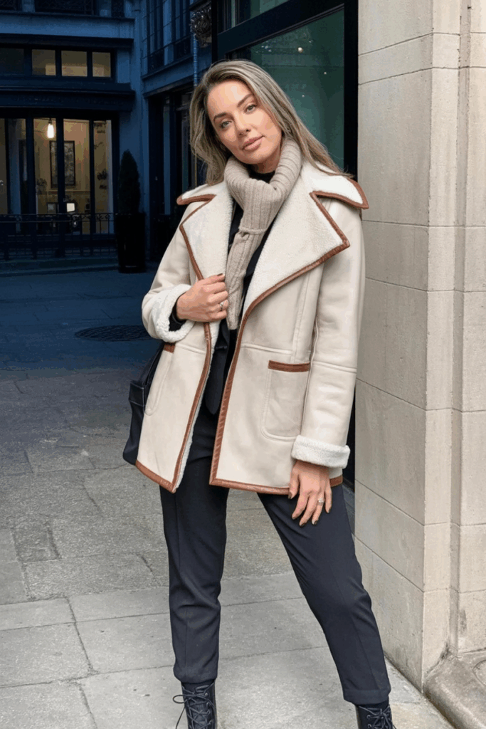 A woman in a cream faux-shearling coat with leather trim, styled with slim trousers, and a chunky scarf