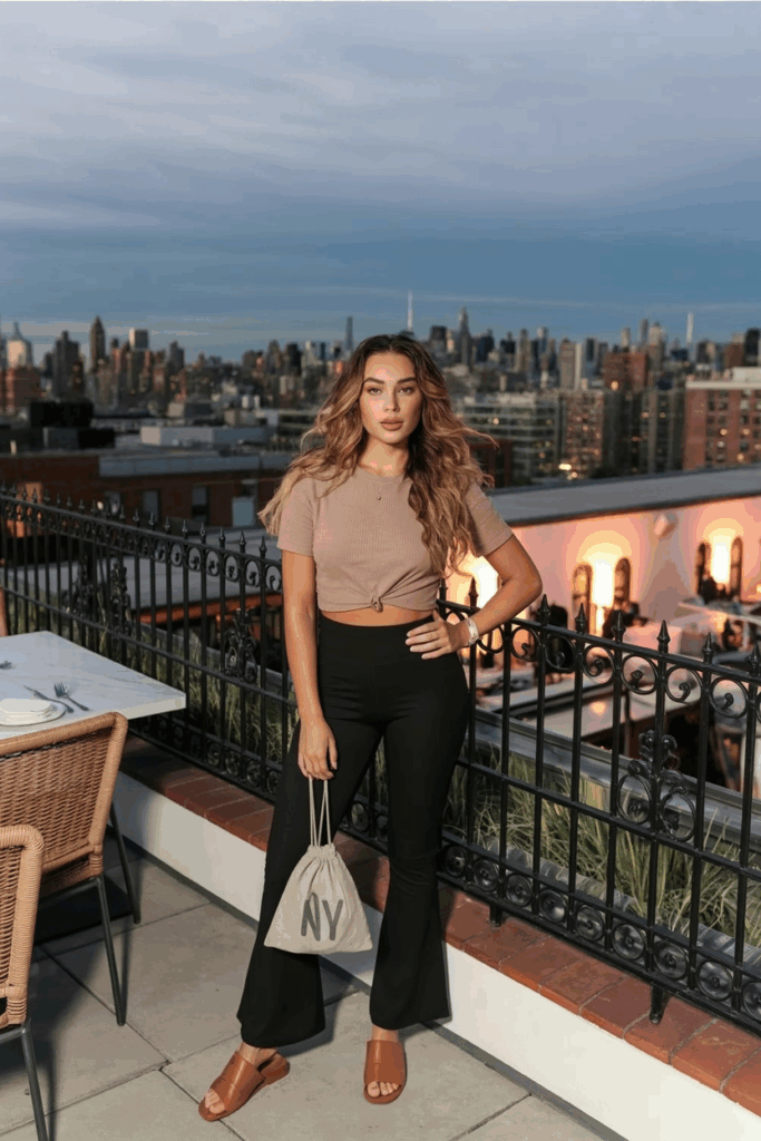 A woman in black flare leggings, a cropped sand-colored boxy tee, brown leather slides, and a drawstring pouch bag.