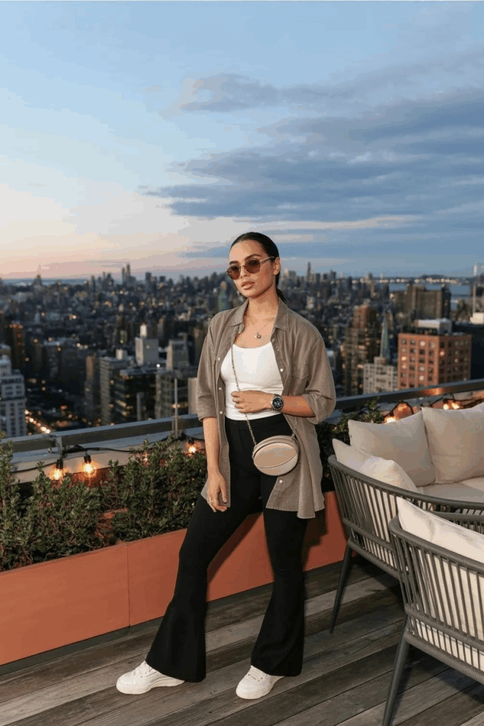 A woman in black flare leggings, a taupe linen button-up shirt layered over a white tank, white leather sneakers, and a crossbody belt bag. She wears aviator sunglasses and a watch