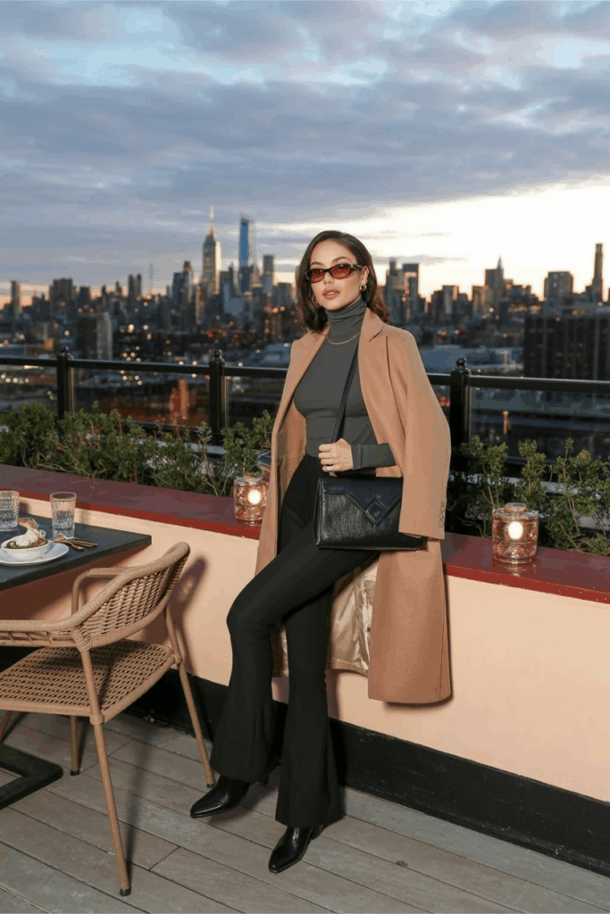 A woman in black flare leggings just grazing the tops of sleek black leather ankle boots with a subtle heel, a fitted charcoal turtleneck, and a longline camel coat. She carries a structured black handbag and wears tortoiseshell sunglasses.