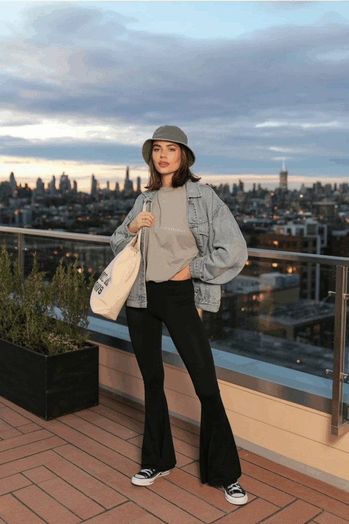 A woman styled in black flare leggings, an oversized light-gray tee, a light-wash denim jacket, and classic white Converse. She wears a bucket hat and carries a reusable tote