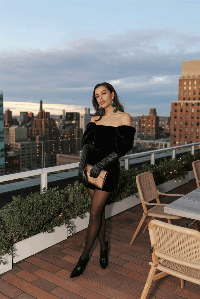 A woman wearing a black velvet dress, black wool-blend tights, emerald earrings, a bronze clutch, leather gloves, and black heeled boot