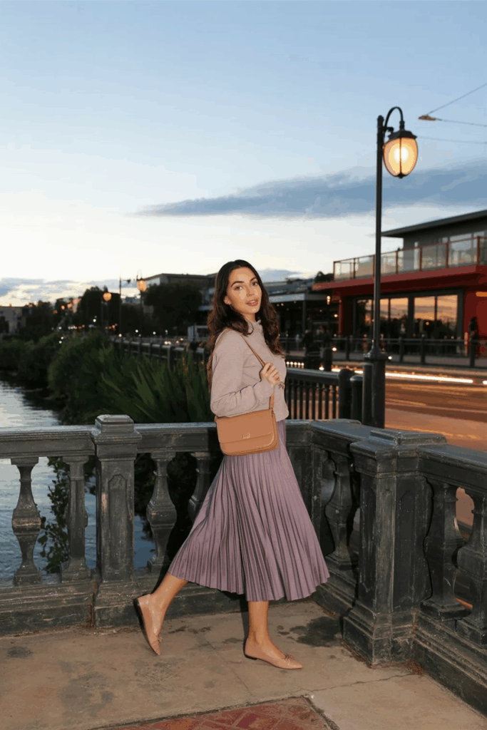 A woman wearing a dusty lavender pleated skirt and nude ballet flats. A soft crossbody bag at her side.