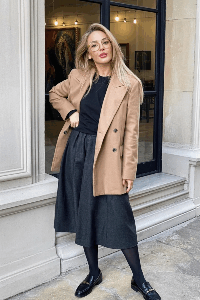 A woman wearing a structured camel double-breasted coat over a charcoal midi skirt and black tights. loafers and gold-rimmed glasses