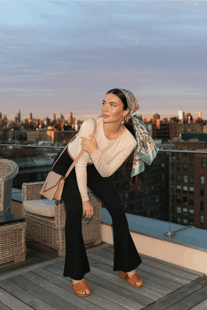 A woman wearing black flare leggings, a soft cream long-sleeve tee, tan leather sandals, and a beige crossbody bag. A printed silk bandana is tied into her low ponytail. She wears thin gold hoop earrings