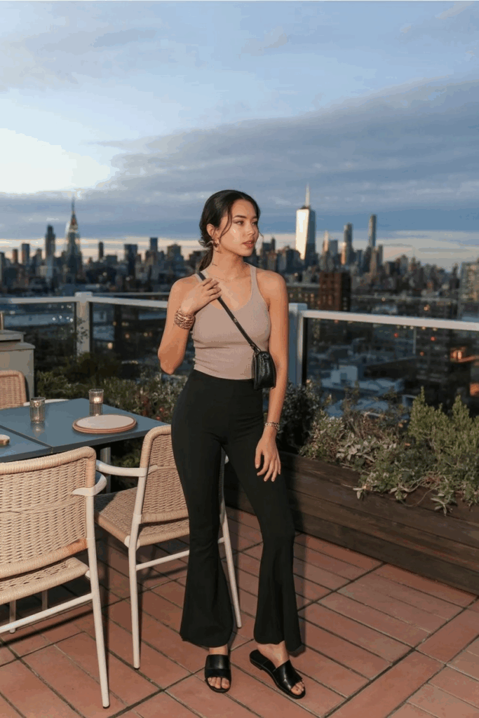 A woman wearing high-waisted black flare leggings, a beige fitted knit top, black leather slide sandals, and a slim crossbody bag. Her hair is loosely tied, and she wears minimal gold jewelry