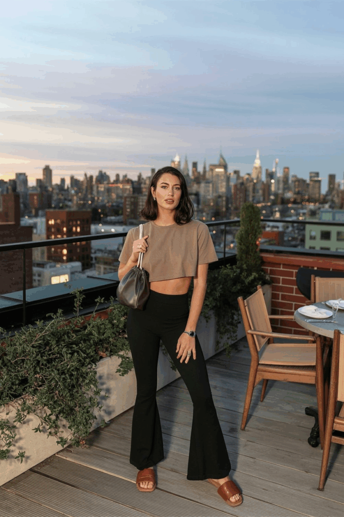 woman in black flare leggings, a cropped sand-colored boxy tee, brown leather slides, and a drawstring pouch bag.
