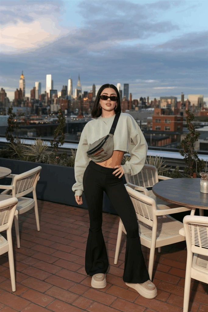 woman in black flare leggings and a pale sage cropped sweatshirt, paired with beige platform sneakers. She accessorizes with a nylon belt bag worn crossbody and oversized sunglasses