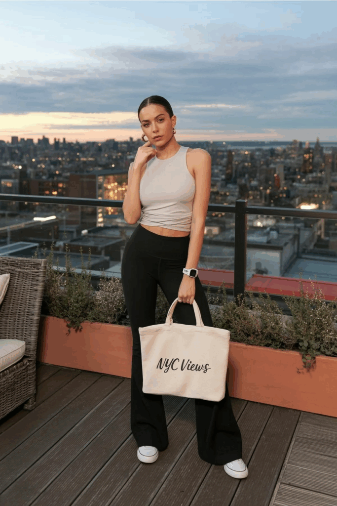 woman in sleek, high-waisted black flare leggings, a tucked-in ivory seamless tank top, white low-top sneakers, and small gold hoops. She carries a canvas tote bag and wears a digital watch