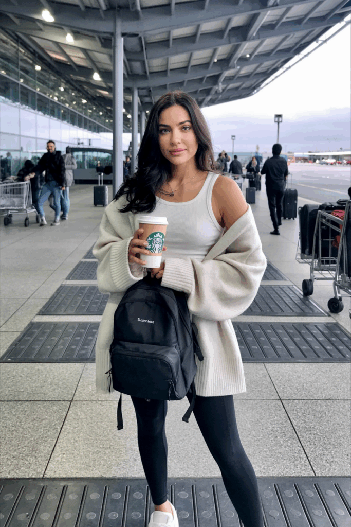 Woman layering a thin white tank top under a cozy oversized cream sweater, paired with stretchy black leggings and simple white trainers, holding a small backpack and a travel coffee cup