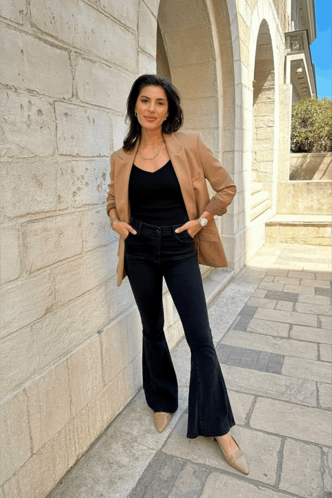 woman wearing black flare leggings, a slim black knit top, a camel blazer, and nude slingback flats.