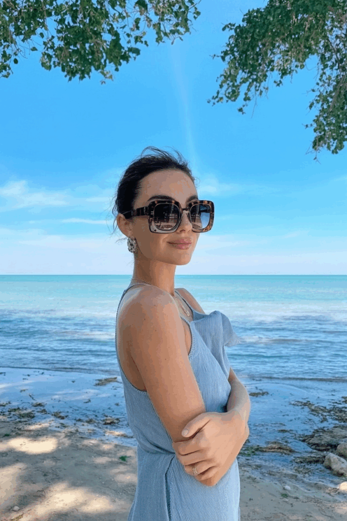 classic tortoiseshell oversized sunglasses, smiling softly, with the ocean blurred in the background.