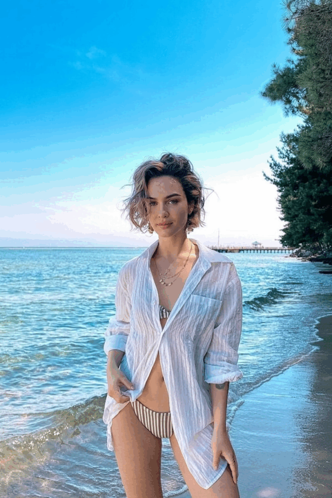 wearing an oversized, sheer white cotton button-down shirt, sleeves rolled, tied loosely at the waist over a striped bikini