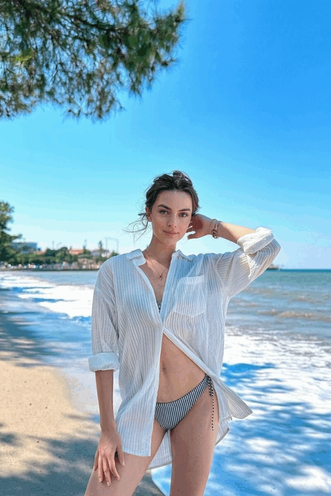 wearing an oversized, sheer white cotton button-down shirt, sleeves rolled, tied loosely at the waist over a striped bikini