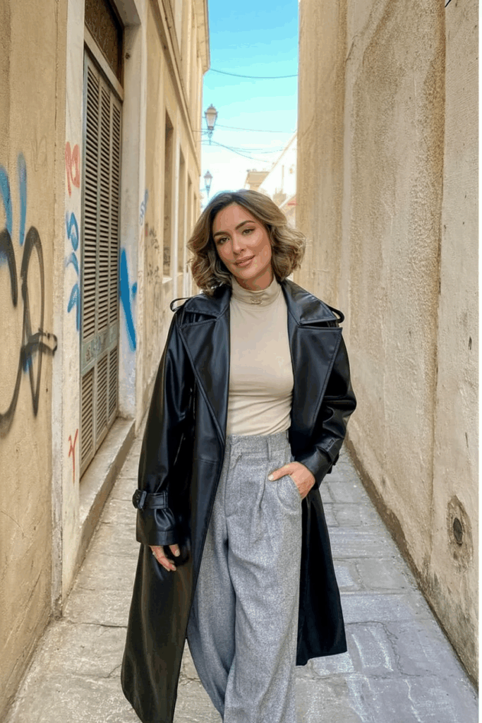 A woman wearing a long black vegan leather trench coat with structured shoulders and a belted waist. Underneath, she wears a soft turtleneck sweater and tapered wool trousers. Her outfit is grounded by sleek mid-heel boots