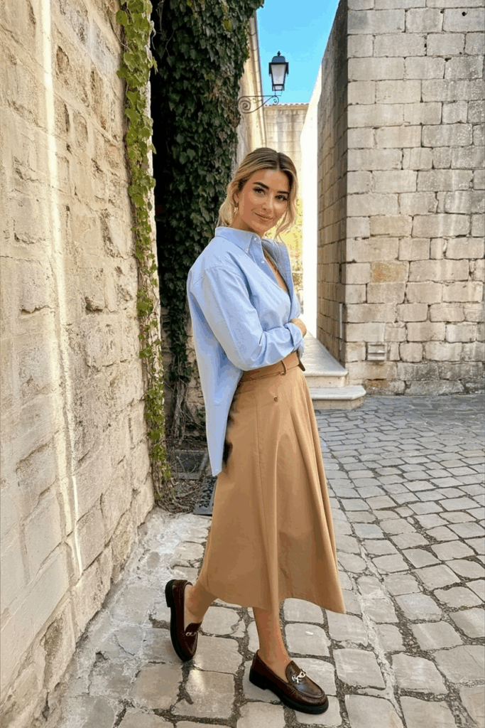 wearing a light blue oxford shirt tucked into a tan midi skirt with chocolate loafers